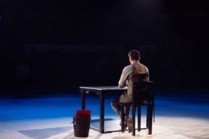 A person sitting alone at a desk under a spotlight in a dark room, symbolizing solitude and contemplation. This image is relevant to discussions on addiction and rehabilitation, such as the support offered by Retorno – Addiction Prevention & Rehabilitation.