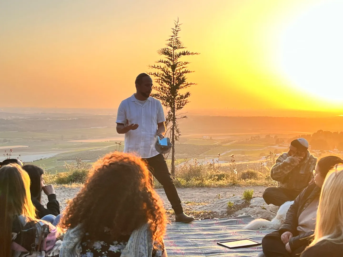 People seated outdoors listening to a speaker at sunset